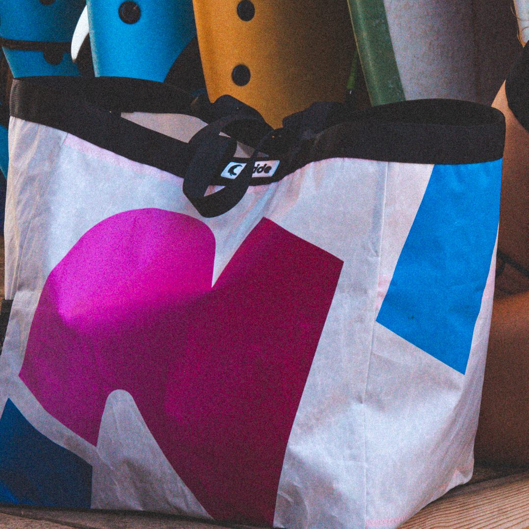 Torba z recyklingu Reride uszyta z żagla kitesurfingowego, w odcieniach różu, bieli i niebieskiego, na tle kolorowych desek surfingowych